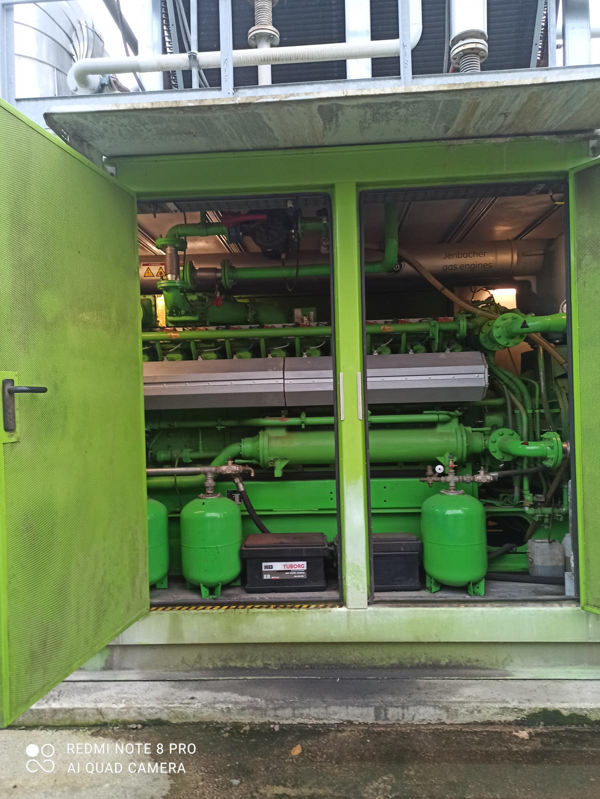 Zielona obudowa z otwartymi drzwiami, wewnątrz widoczny silnik Jenbacher gaz engine i elementy instalacji.